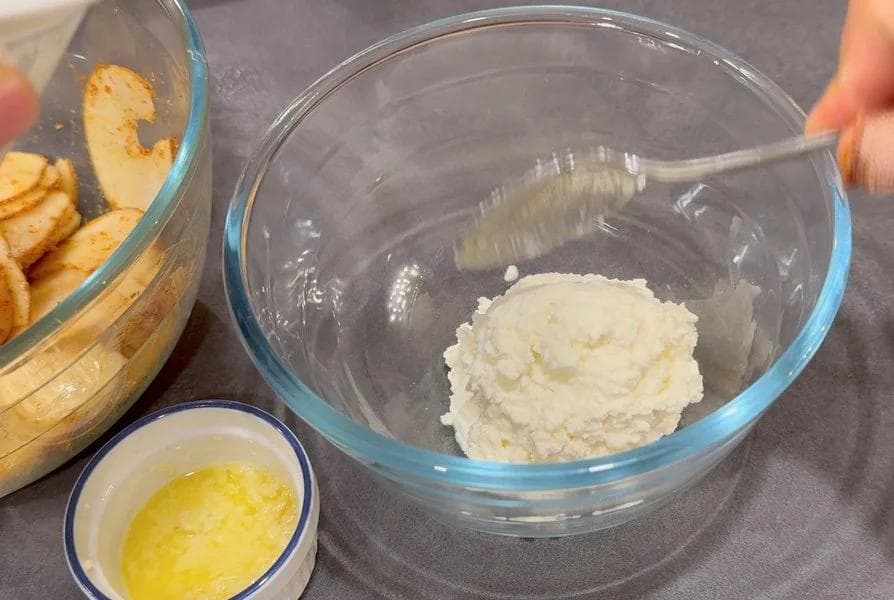 Ricotta mixture combined with maple syrup and lime juice in a glass bowl with a spoon, beside sliced potatoes and melted garlic butter.