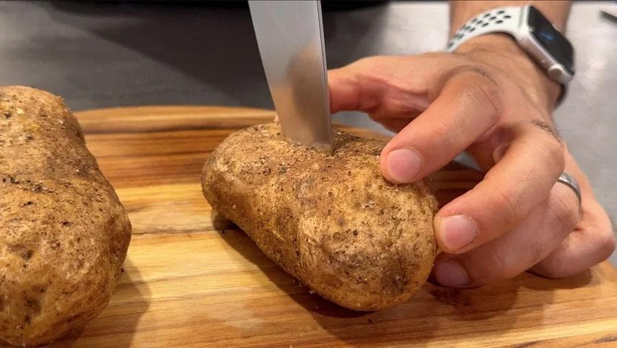 Hand holding a halved potato on a wooden cutting board while slicing it horizontally with a knife.