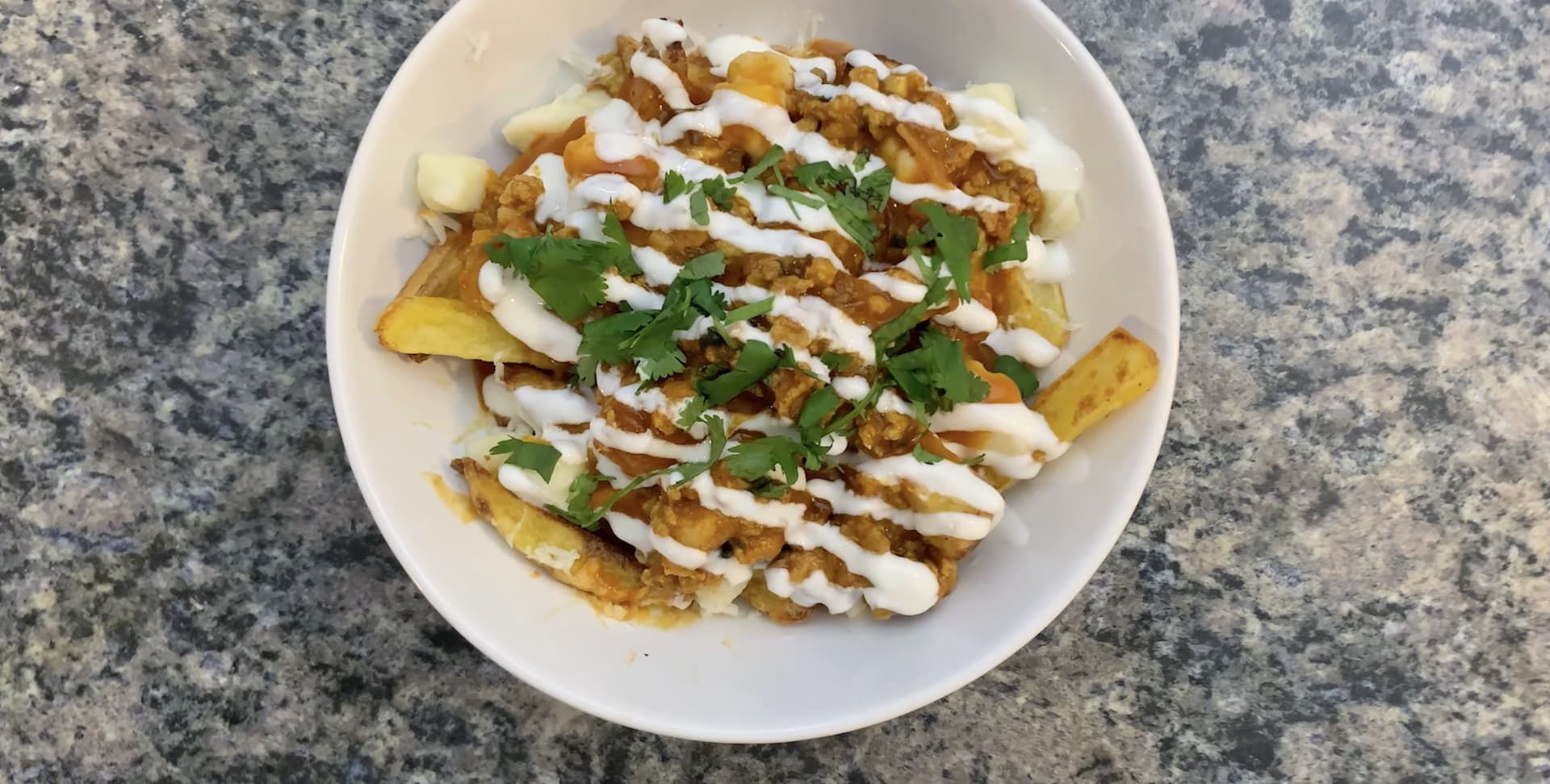 Indian Fusion Aloo Keema Poutine with crispy fries, meat sauce, creamy topping, cilantro and green onions on white plate.