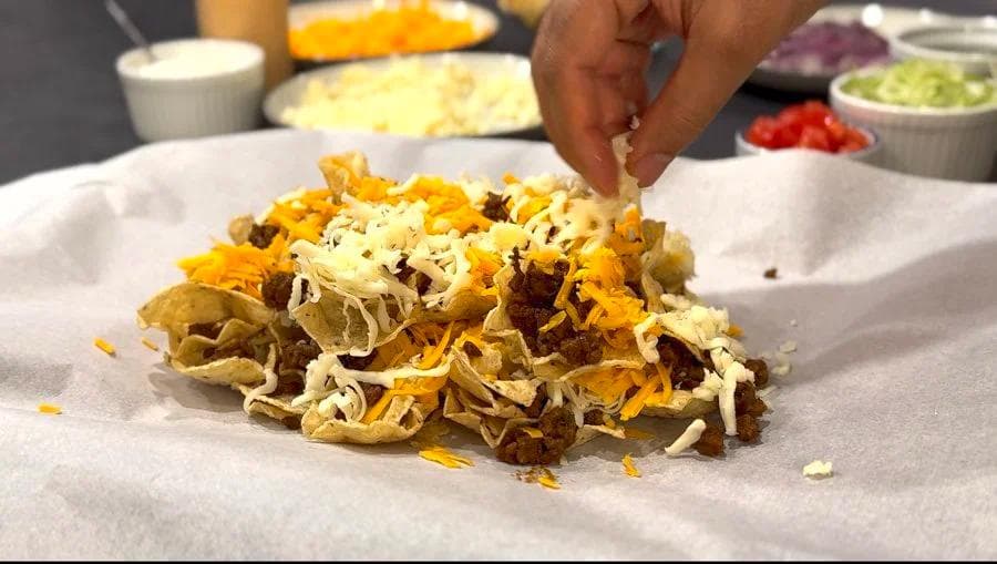Hand adding shredded cheese to a pile of crispy tortilla chips and ground beef on parchment paper.
