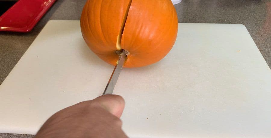 Cutting a whole pumpkin in half with a knife around the stem on parchment paper.