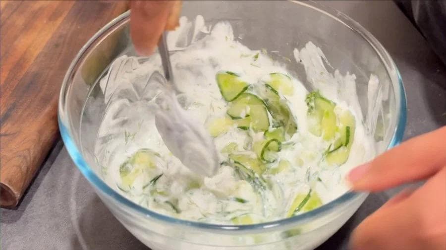 Mixing yogurt-based dressing with sliced cucumbers and dill in a glass bowl using a spatula.
