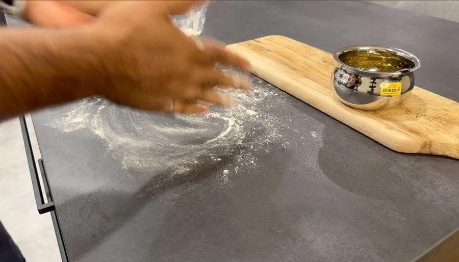 Hand rolling out palak paratha dough on a dark surface with flour, wooden rolling pin and ghee nearby.