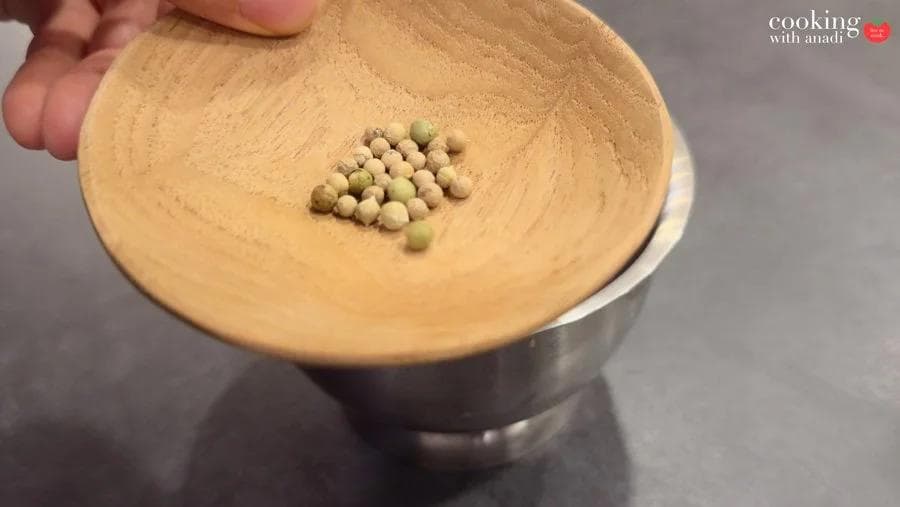 White peppercorns in a small wooden bowl held over a stainless steel mortar, ready to be crushed.