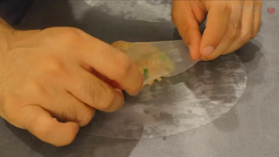 Hands folding a rice paper wrapper around chicken filling into a rectangular dumpling shape on a gray surface.