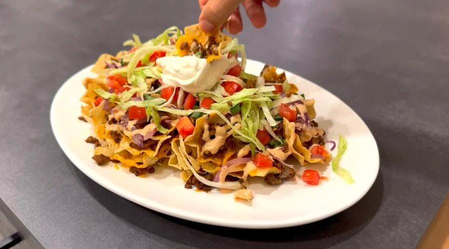 Cheeseburger nachos on a white plate topped with sour cream, shredded lettuce, diced tomatoes, and melted cheese.