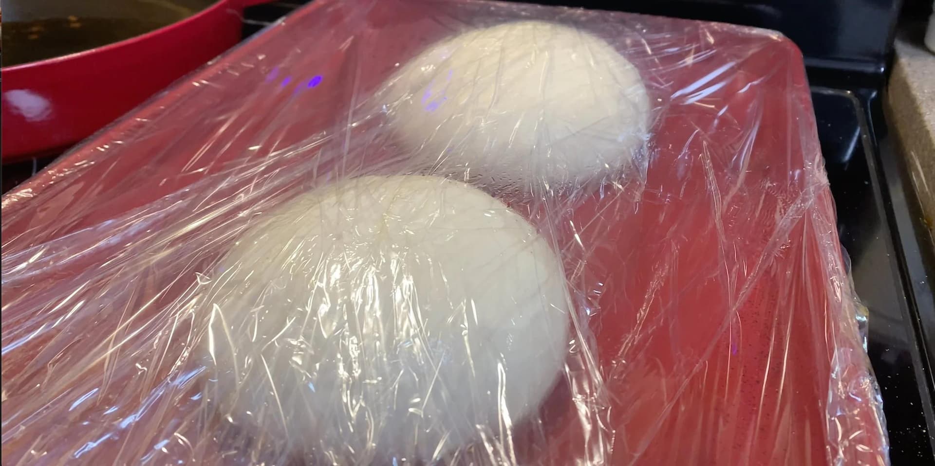 Two portions of pizza dough resting on a red tray, covered with plastic wrap.