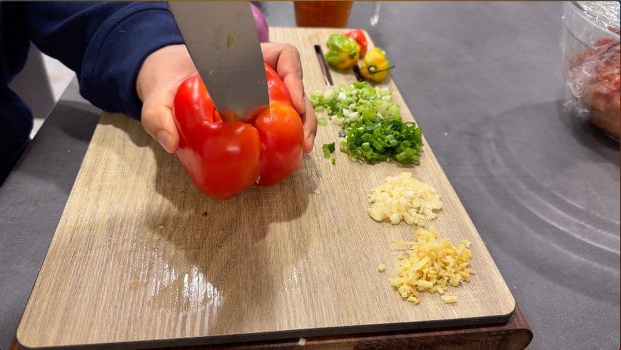 Hands holding red bell peppers over a cutting board with chopped garlic, ginger, and green onions for Jamaican pepper steak preparation.