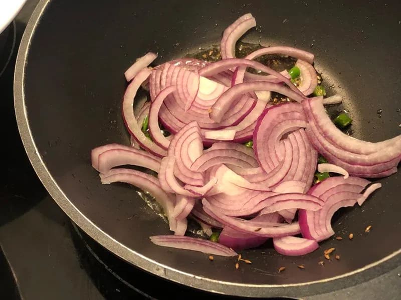 Sliced red onions and chopped green chilies cooking in oil in a dark pan