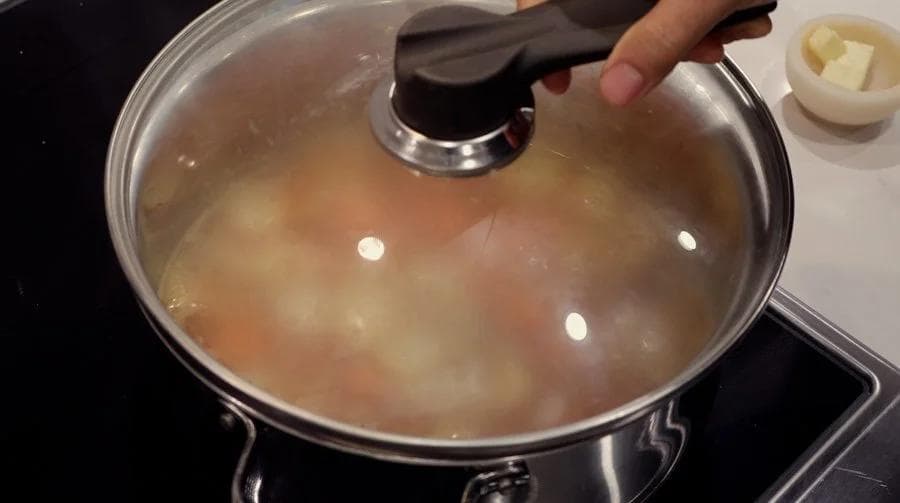 Pot of simmering carrot and ginger soup covered with a glass lid on the stovetop.