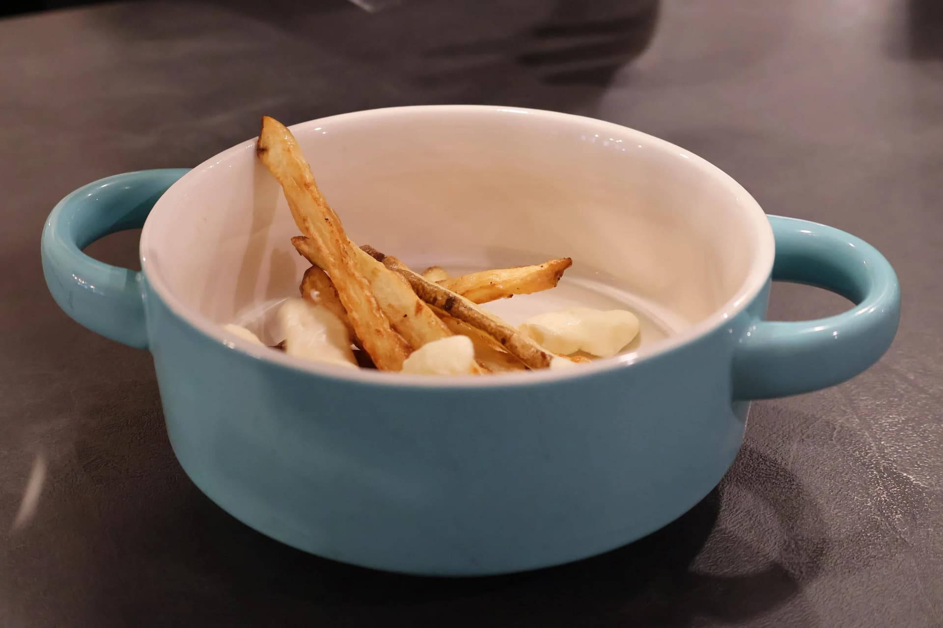Crispy fries and cheese curds in a blue bowl with gravy being poured over them for poutine assembly.