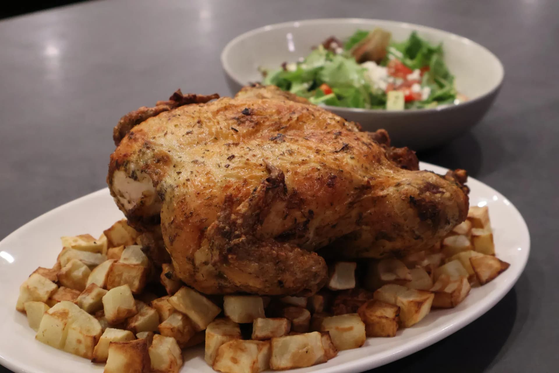 Whole air fryer chicken on a white oval platter surrounded by golden diced potatoes, with a green salad in a bowl in the background.