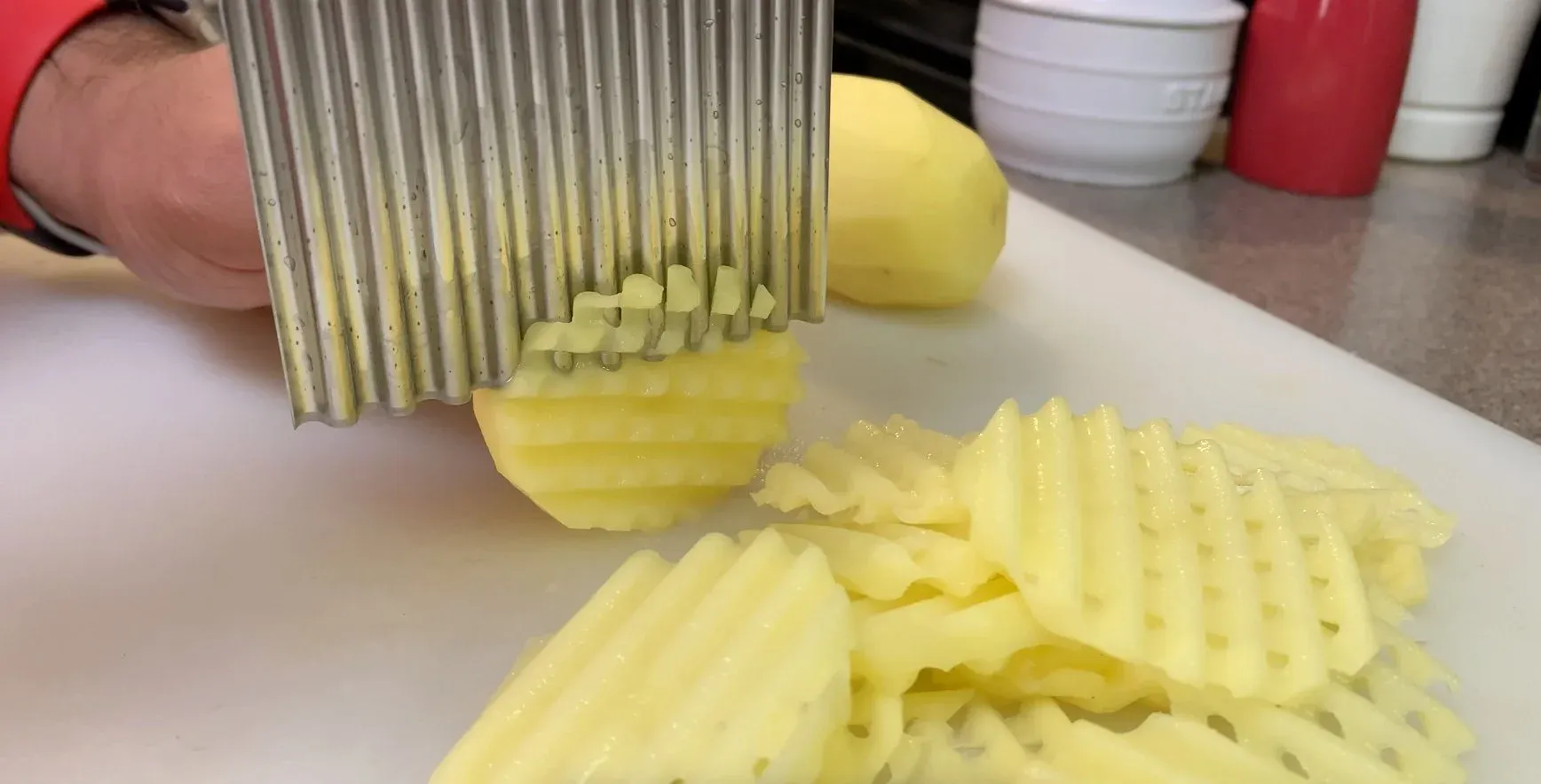 Crinkle cutter slicing a potato lengthwise to create waffle fry cuts on a white surface.