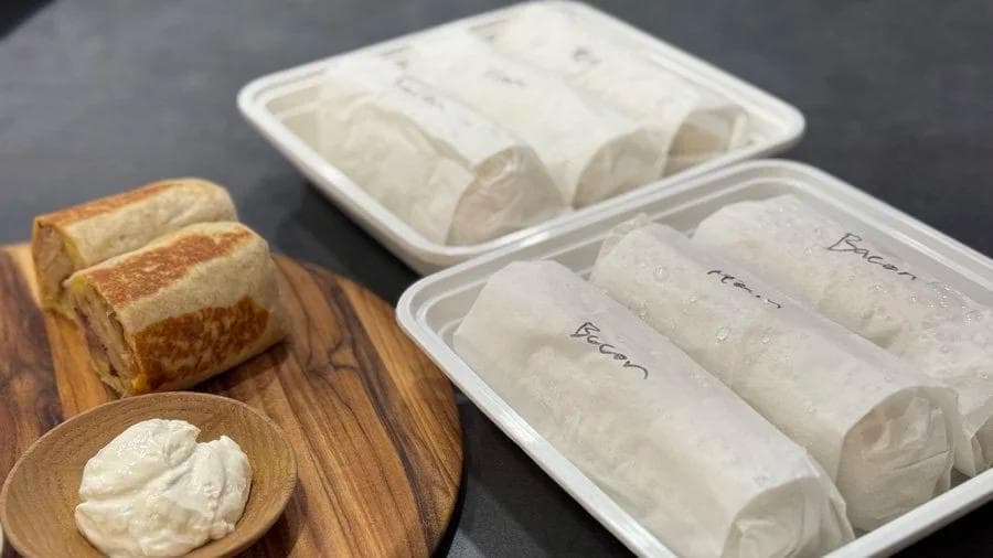 Meal prep breakfast burritos wrapped in paper with labels, served with toasted bread and cream cheese on a wooden board.