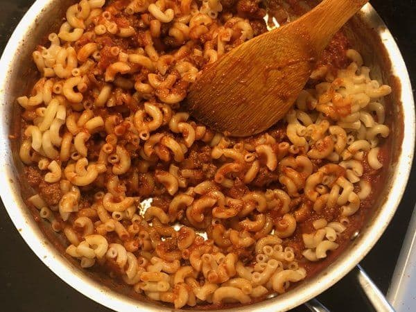 Elbow macaroni mixed with seasoned ground beef and tomato-based sauce in a white pot with wooden spoon.