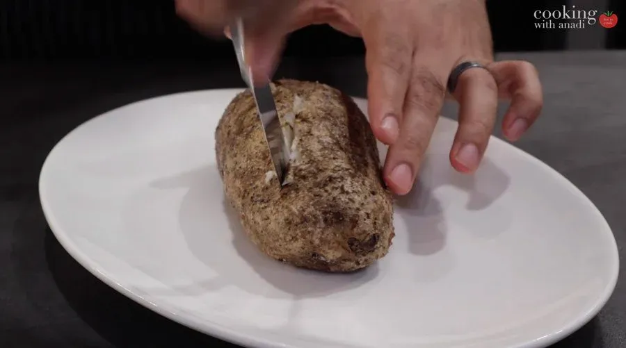 Slicing open a baked potato in a cross pattern on a white plate, with hands pushing the ends to fluff the interior.