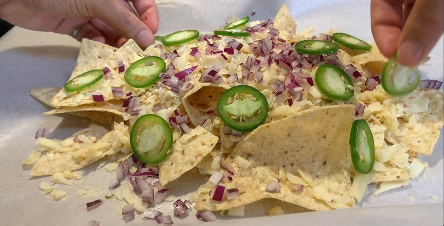 Buffalo chicken nachos topped with melted cheese, sliced jalapeños, and diced red onions on a white plate.