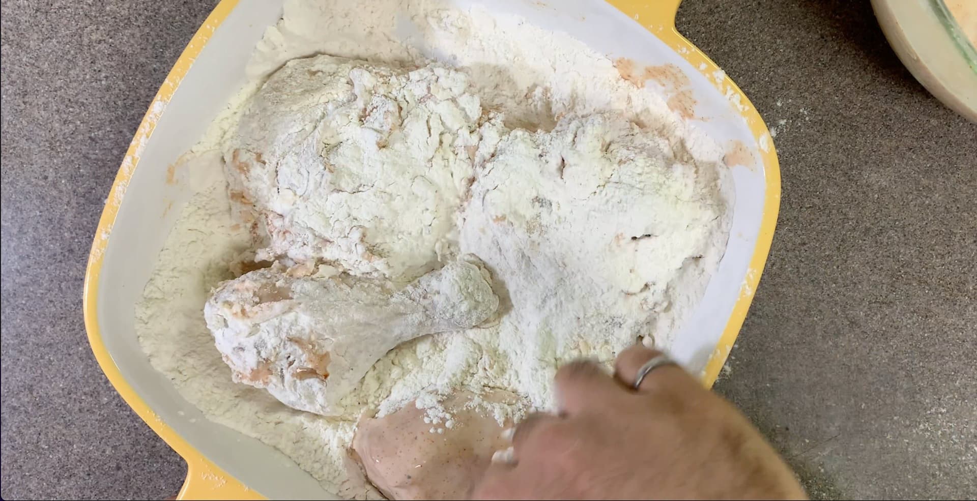 Hand coating raw chicken pieces in seasoned flour mixture on a yellow-rimmed white plate.