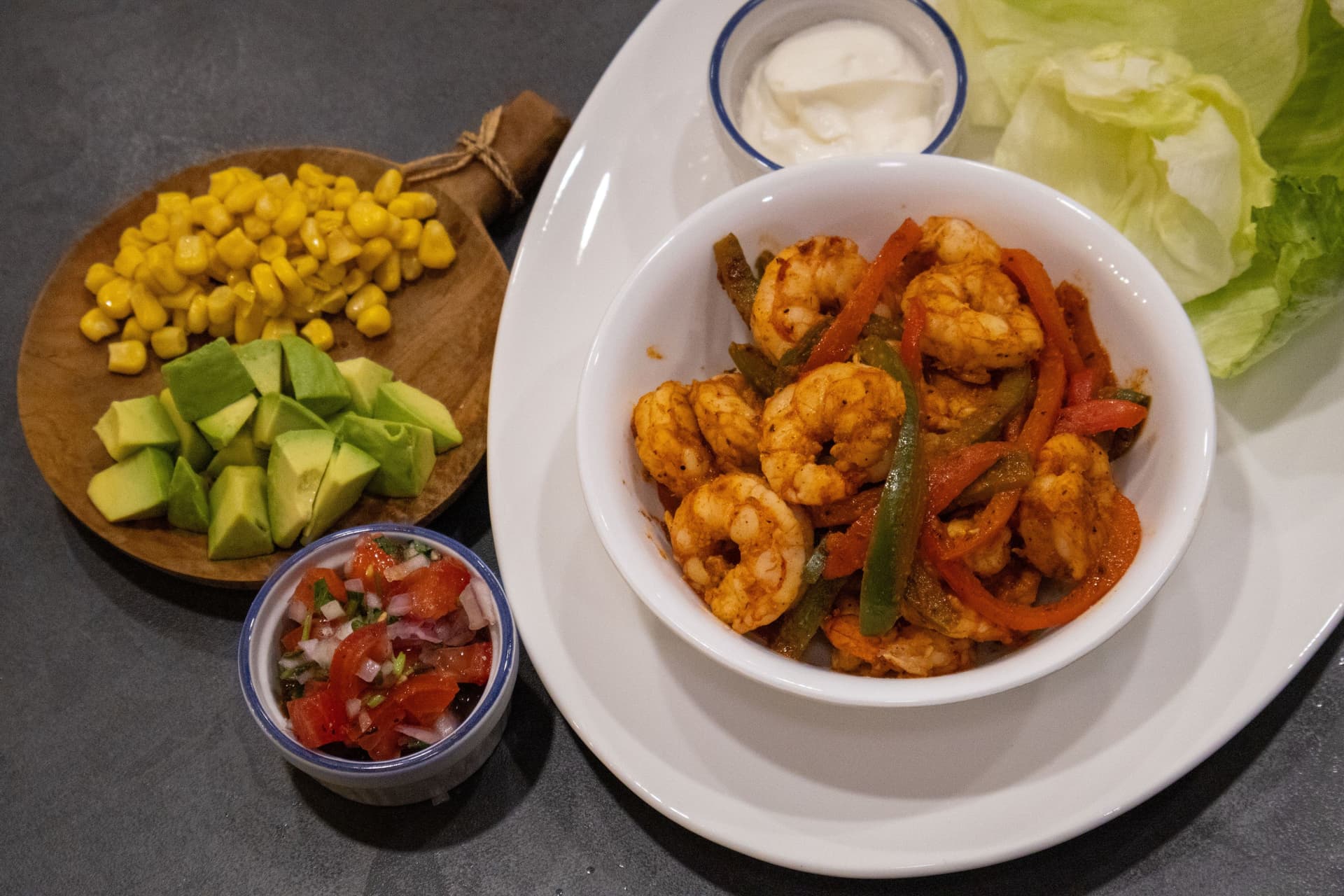 Cajun-spiced shrimp with peppers in a white bowl, surrounded by corn, avocado, salsa, sour cream, and lettuce wraps.