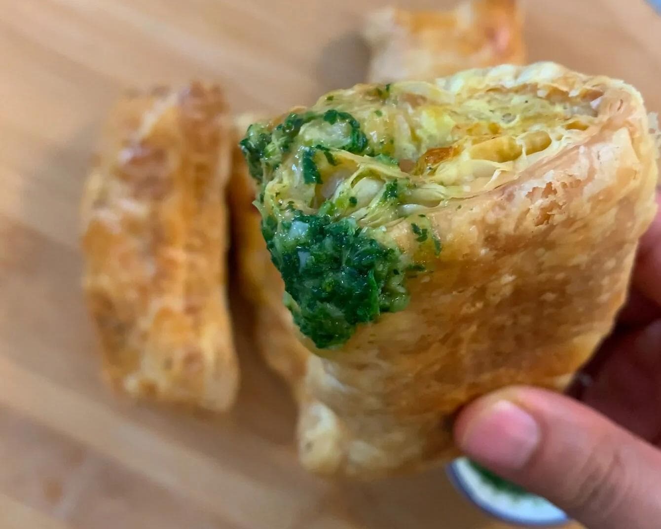 Paneer puff patties held in hand, showing golden pastry with green paneer and herb filling visible inside.