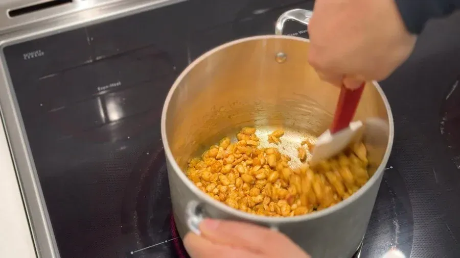 Stirring roasted peanuts and spices in a pot with a red spatula while preparing peanut sauce.