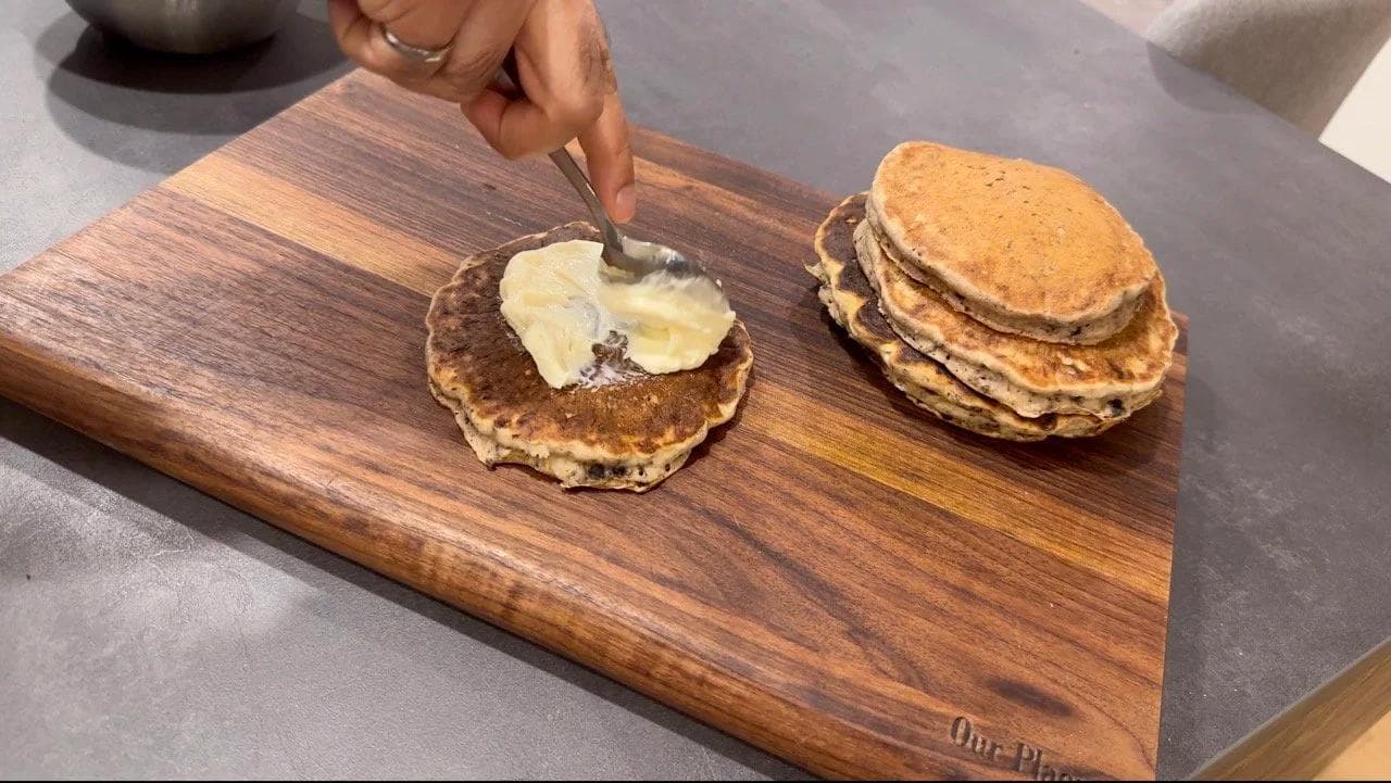 Assembling gluten-free Oreo cheesecake pancakes by spreading cream cheese glaze between stacked pancakes on a wooden board.