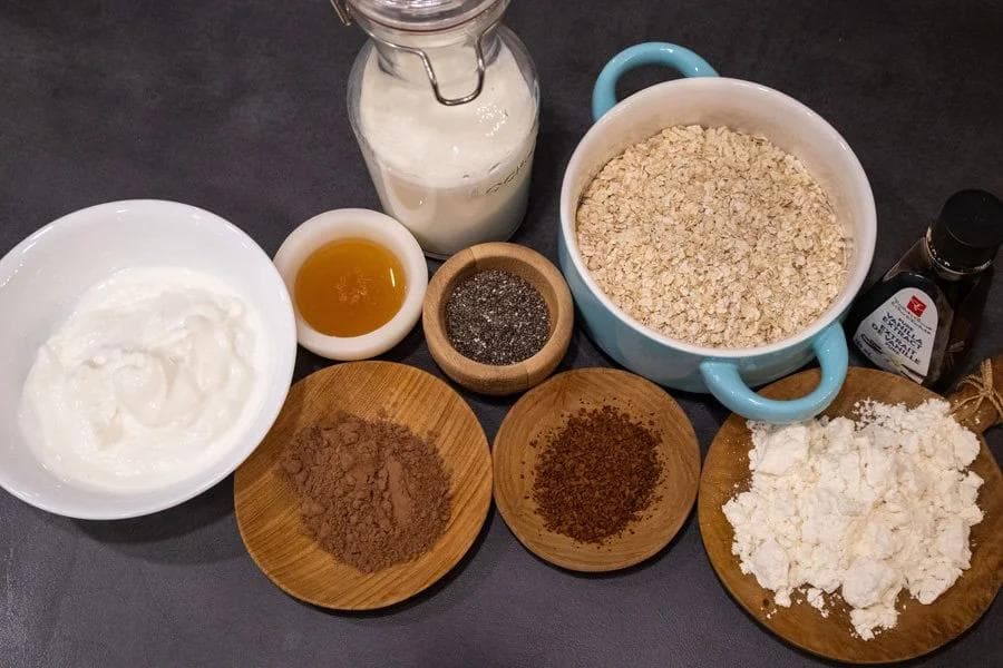 Ingredients for tiramisu overnight oats including milk, yogurt, oats, honey, cocoa powder, and cottage cheese on a dark surface.