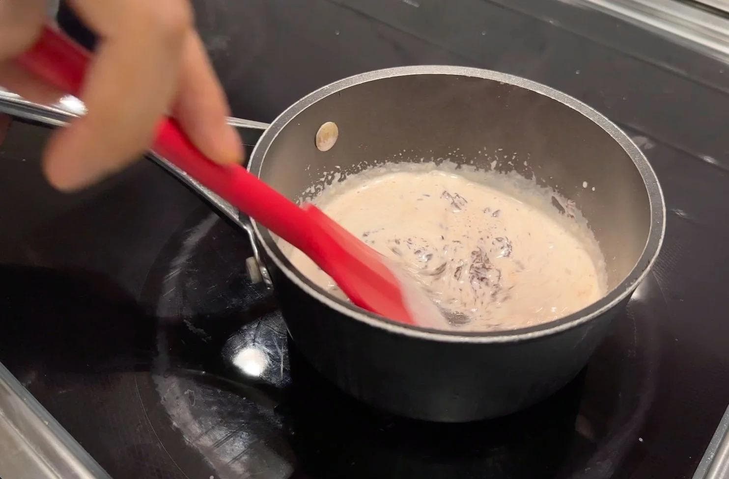Stirring chocolate chips into steaming cream in a saucepan with a red spatula.