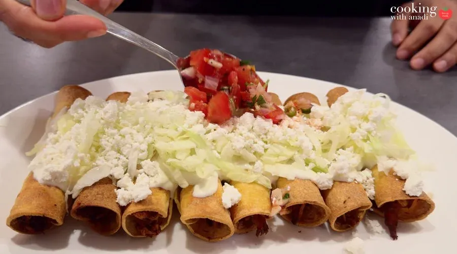 Taquitos arranged on a white plate, topped with shredded lettuce, crumbled cotija, crema, and pico de gallo being spooned on top.