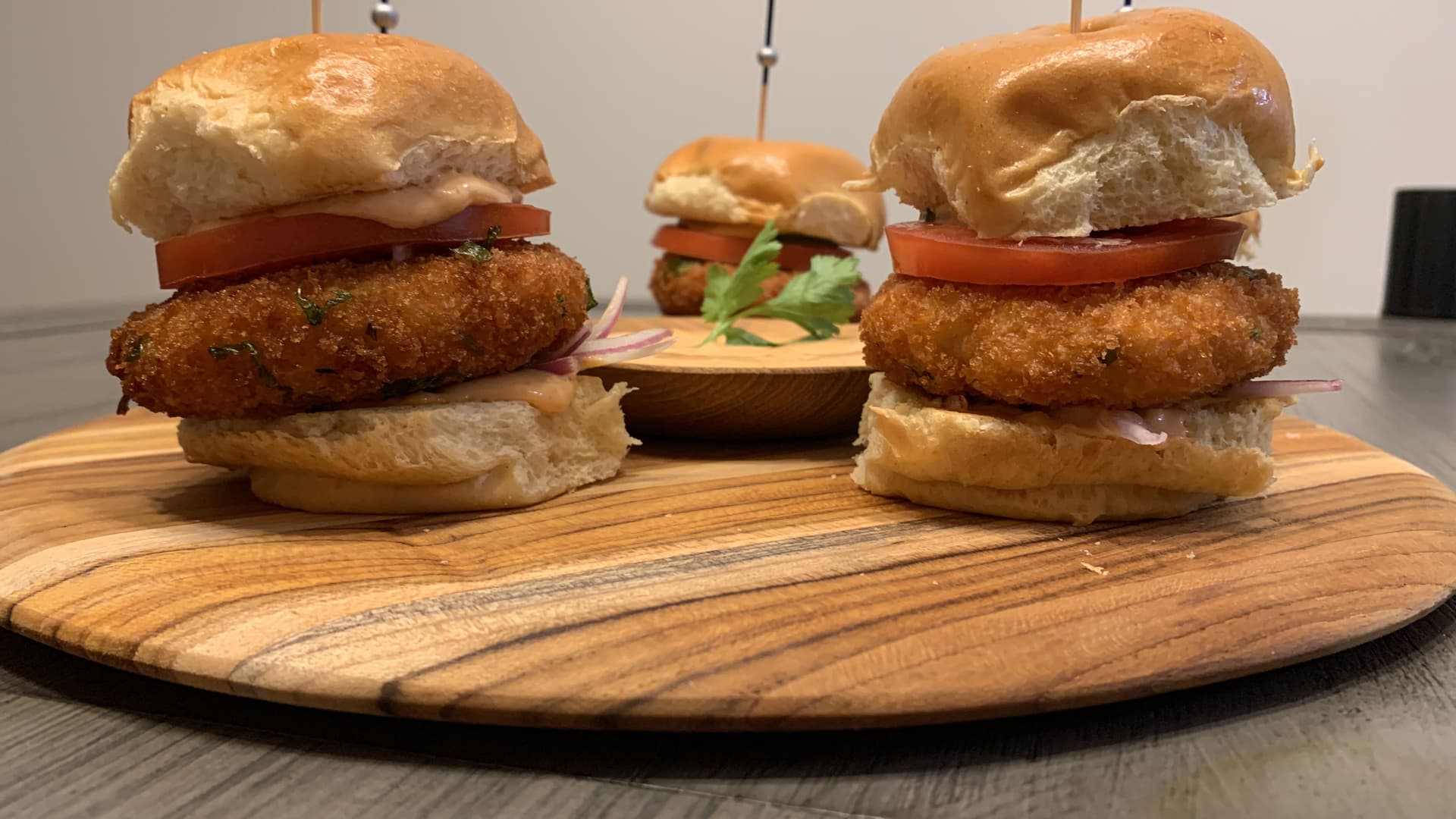 Three crispy golden fried potato patty burgers on toasted buns with tomato slices, arranged on a wooden board with a small dipping sauce.