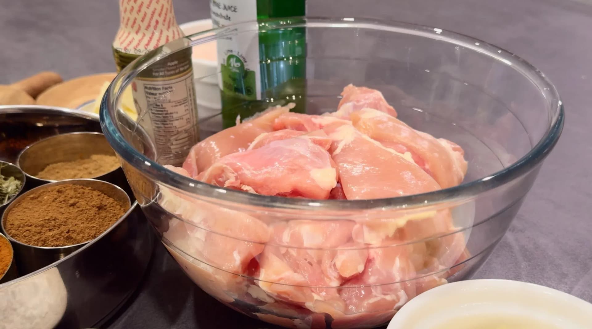 Raw chicken thighs in a large glass bowl after first marinade, surrounded by small bowls of spices and seasonings.