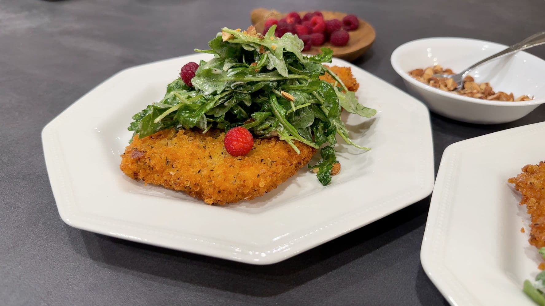 Panko-crusted chicken schnitzel topped with fresh arugula and raspberries on a white plate.