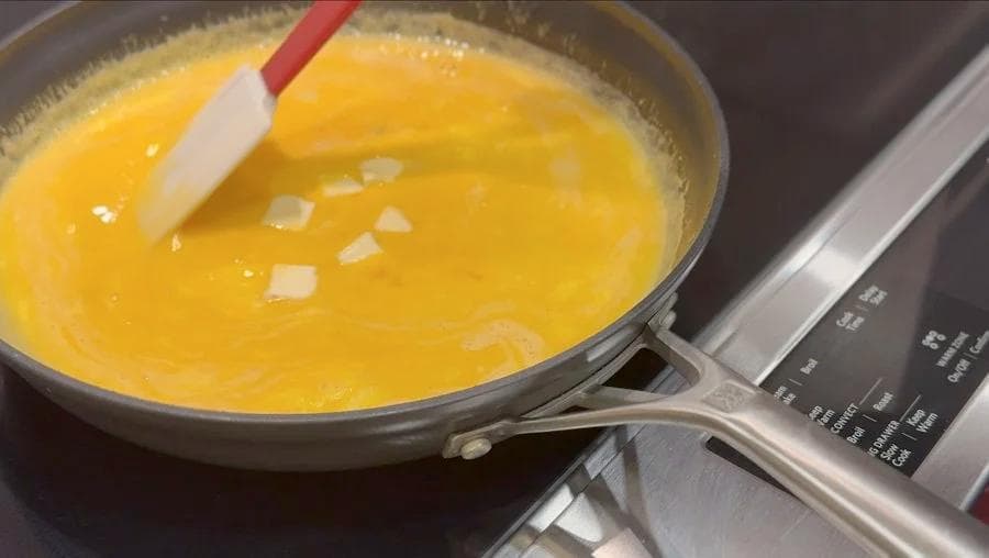 Whisking scrambled eggs in a skillet with diced onions visible on the surface, using a red and white spatula.