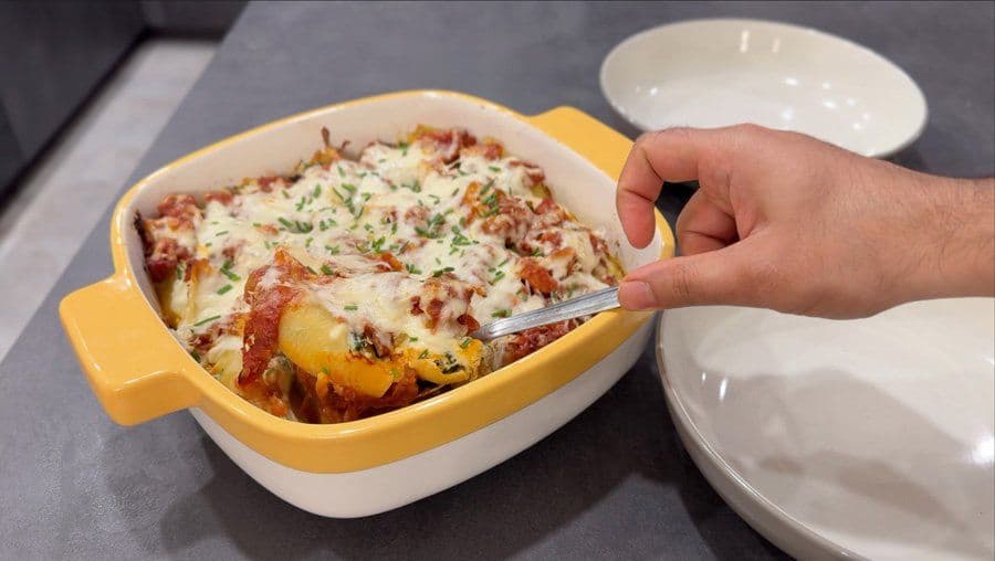 Cheesy stuffed shells with amatriciana sauce in a yellow and white baking dish, garnished with chives, being served with a spoon.