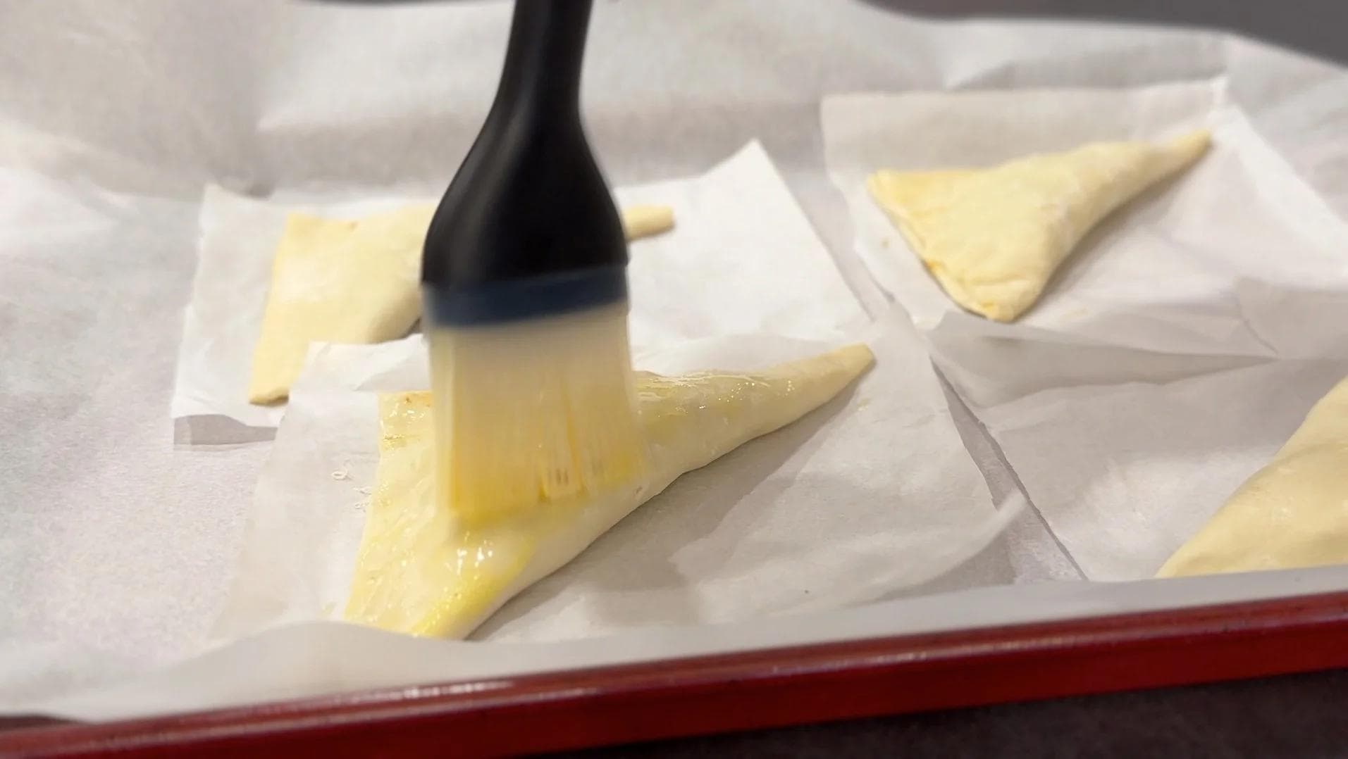 Brushing egg wash onto folded puff pastry triangles with a black pastry brush on parchment paper.