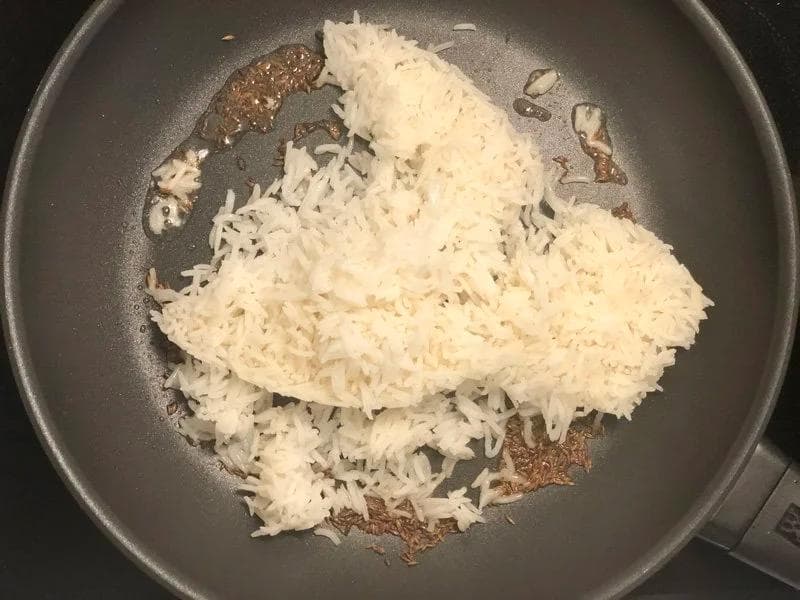 Shredded white rice being stirred in a dark pan with crackled cumin seeds and oil.