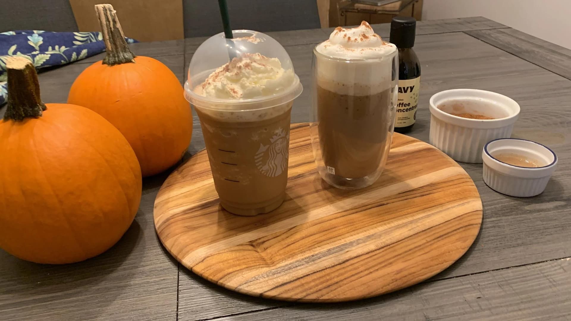 Homemade pumpkin spice frappuccinos topped with whipped cream, served in glasses alongside fresh pumpkins and coffee ingredients on a wooden board.