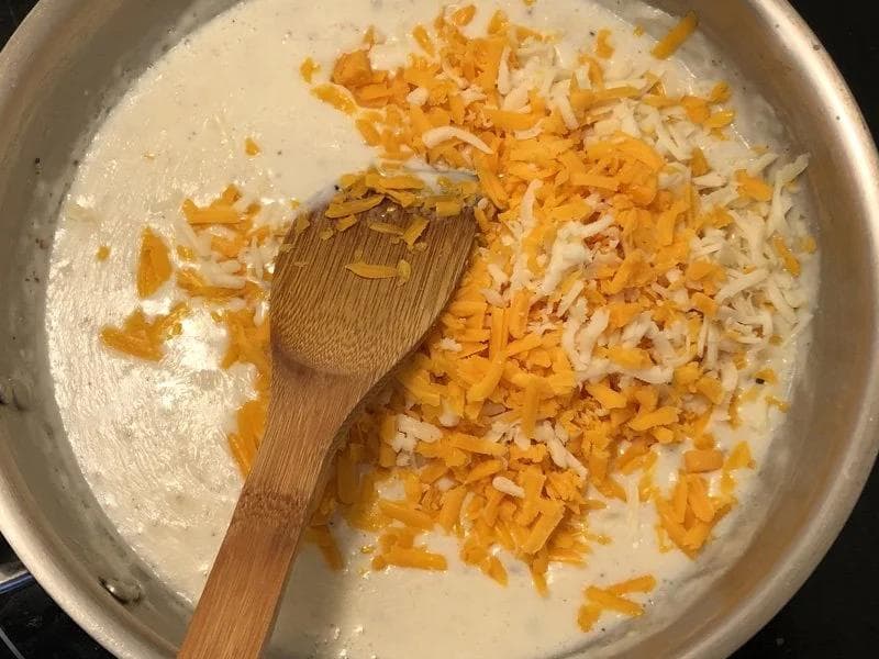 Wooden spoon stirring shredded cheddar and mozzarella cheese into a creamy béchamel sauce in a stainless steel pot.