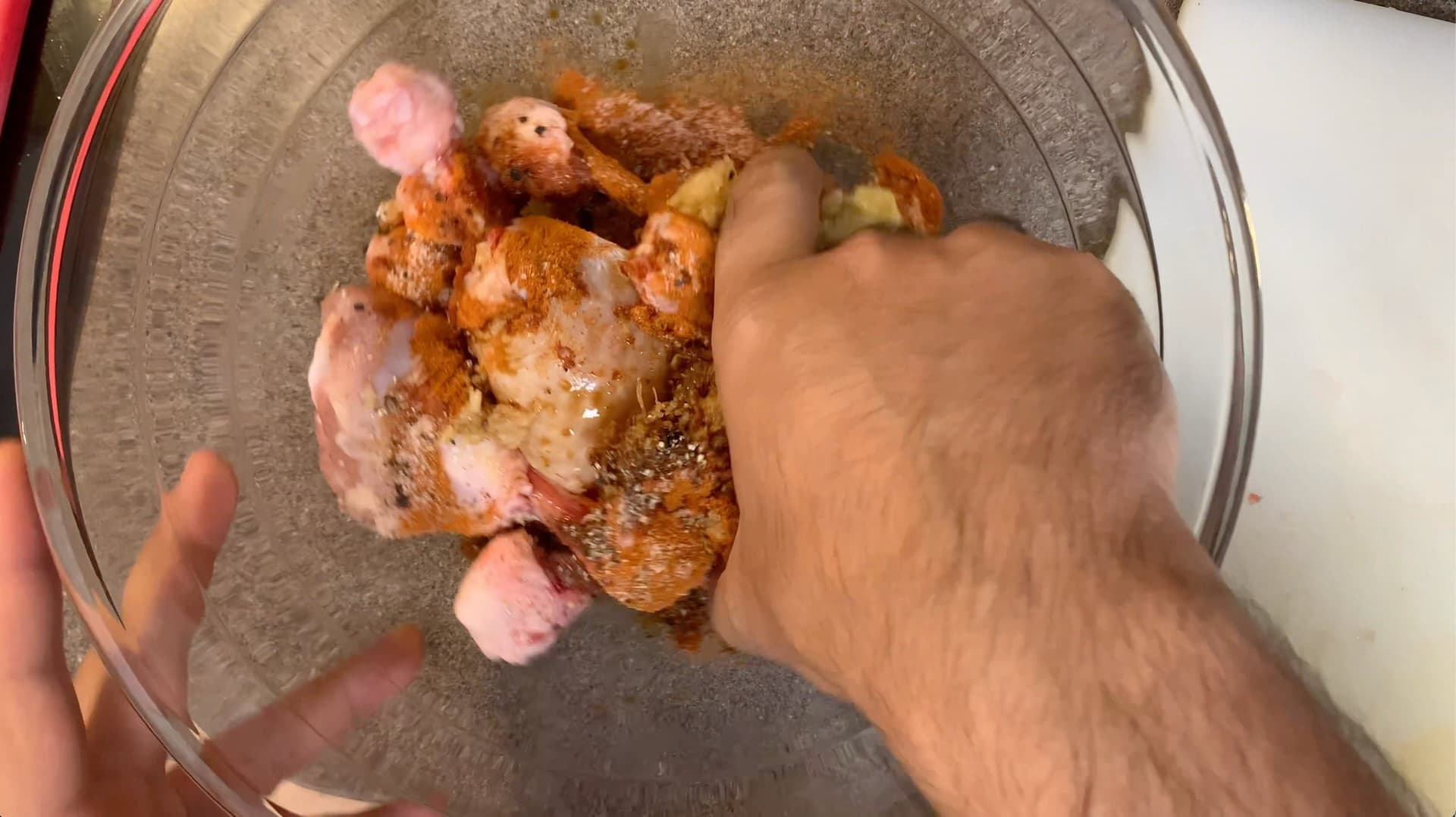 Marinated chicken drumsticks being tossed in a glass bowl with spices and seasonings.