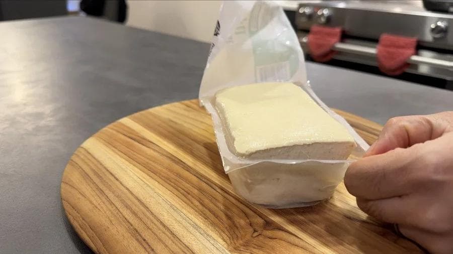Hand holding package of tofu over wooden cutting board during prep step.