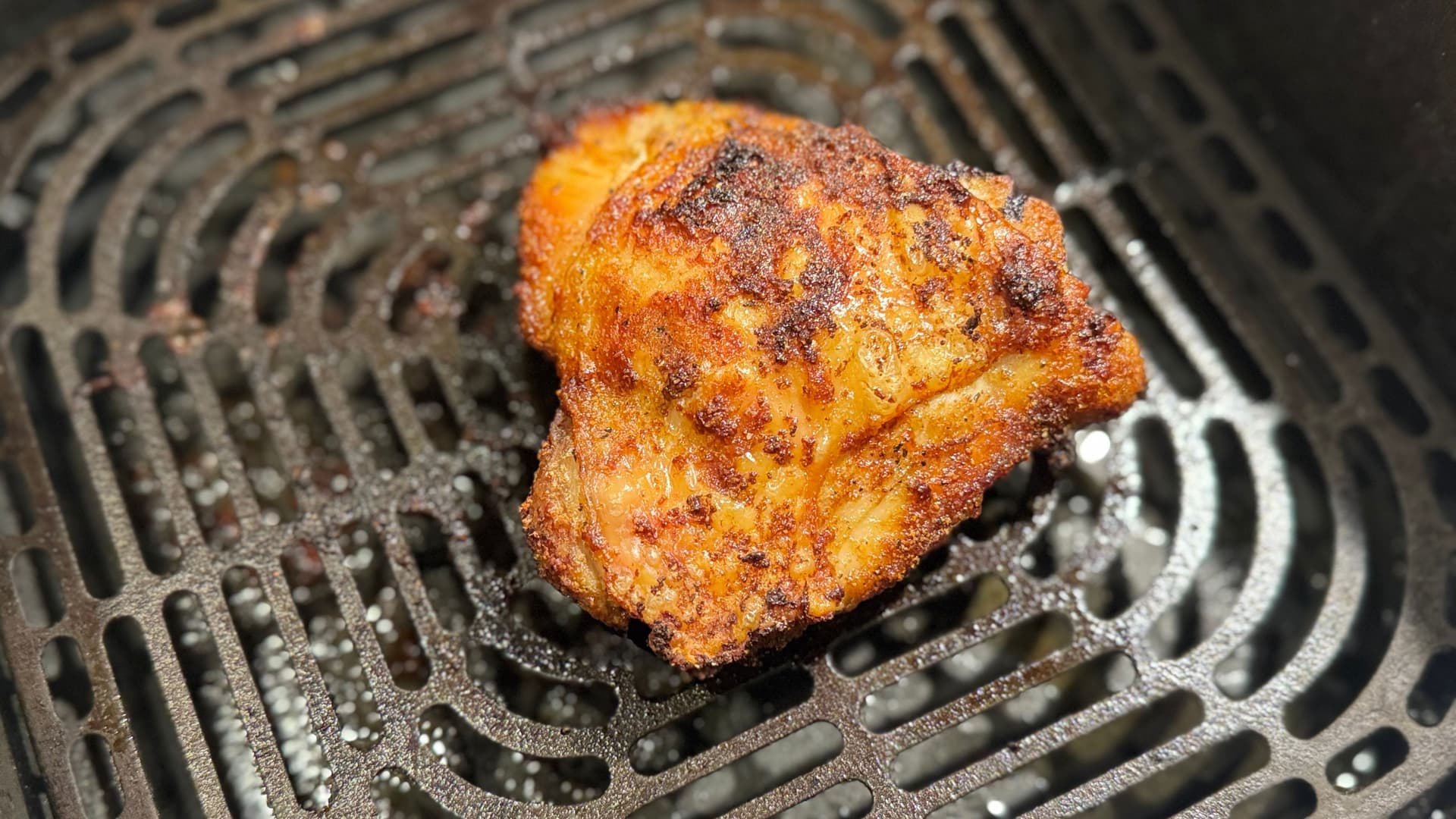Golden-brown crispy chicken thigh cooking on a grill grate with seasoning visible on the skin.