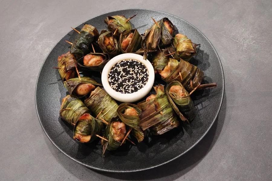 Pandan leaf-wrapped chicken parcels arranged on a dark plate around a small bowl of black sesame dipping sauce.