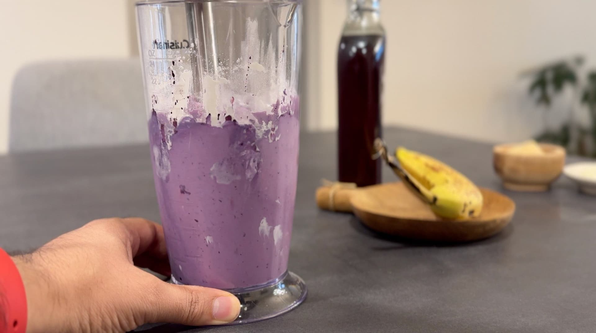 Hand holding a glass of purple blueberry cheesecake protein ice cream with a banana and dark syrup bottle in the background.