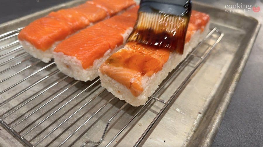 Three pieces of oshizushi with salmon being brushed with teriyaki reduction on a wire rack over a sheet pan.