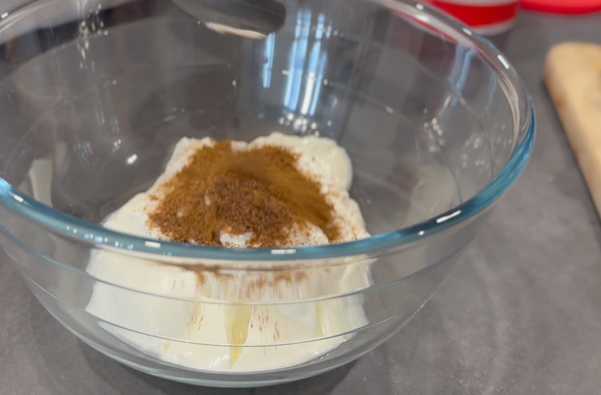 Yogurt with ground cumin powder sprinkled on top in a glass mixing bowl, ready to be whisked together.