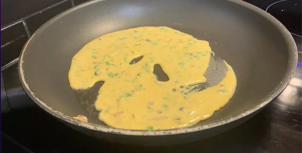 Besan chilla cooking in a pan with ghee, showing the yellow savory pancake with herbs and holes forming as it sets.