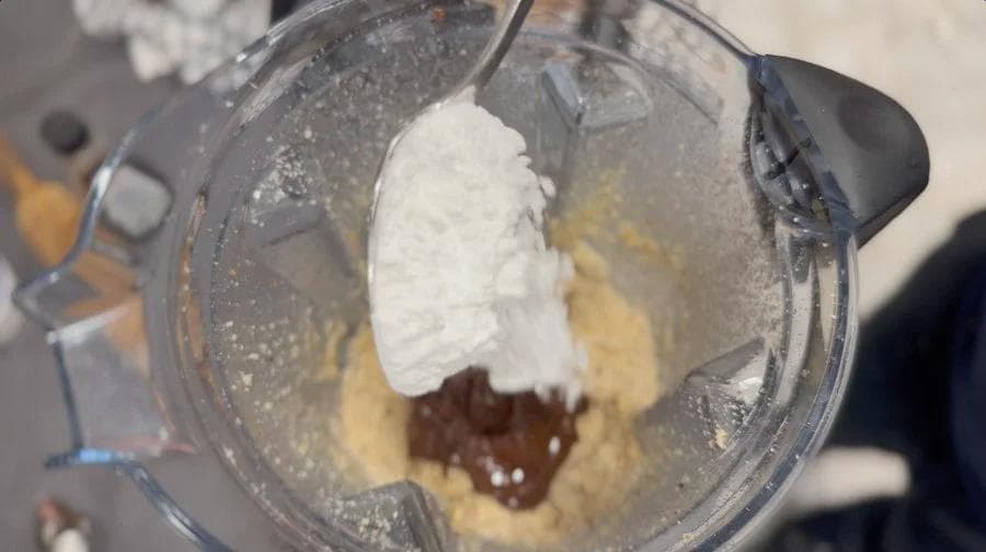 Food processor bowl with melted chocolate, white powder, and brown mixture being blended into vegan Nutella.