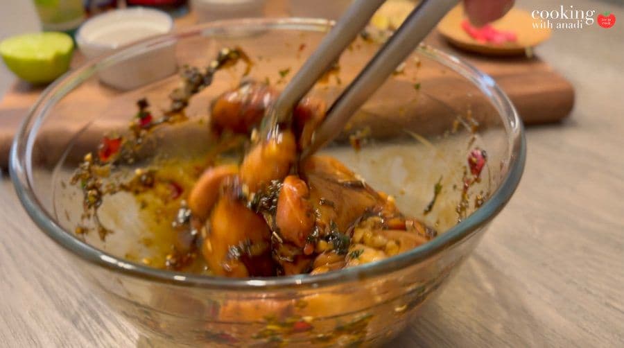 Marinated chicken with herbs and spices mixed in a glass bowl with metal tongs.