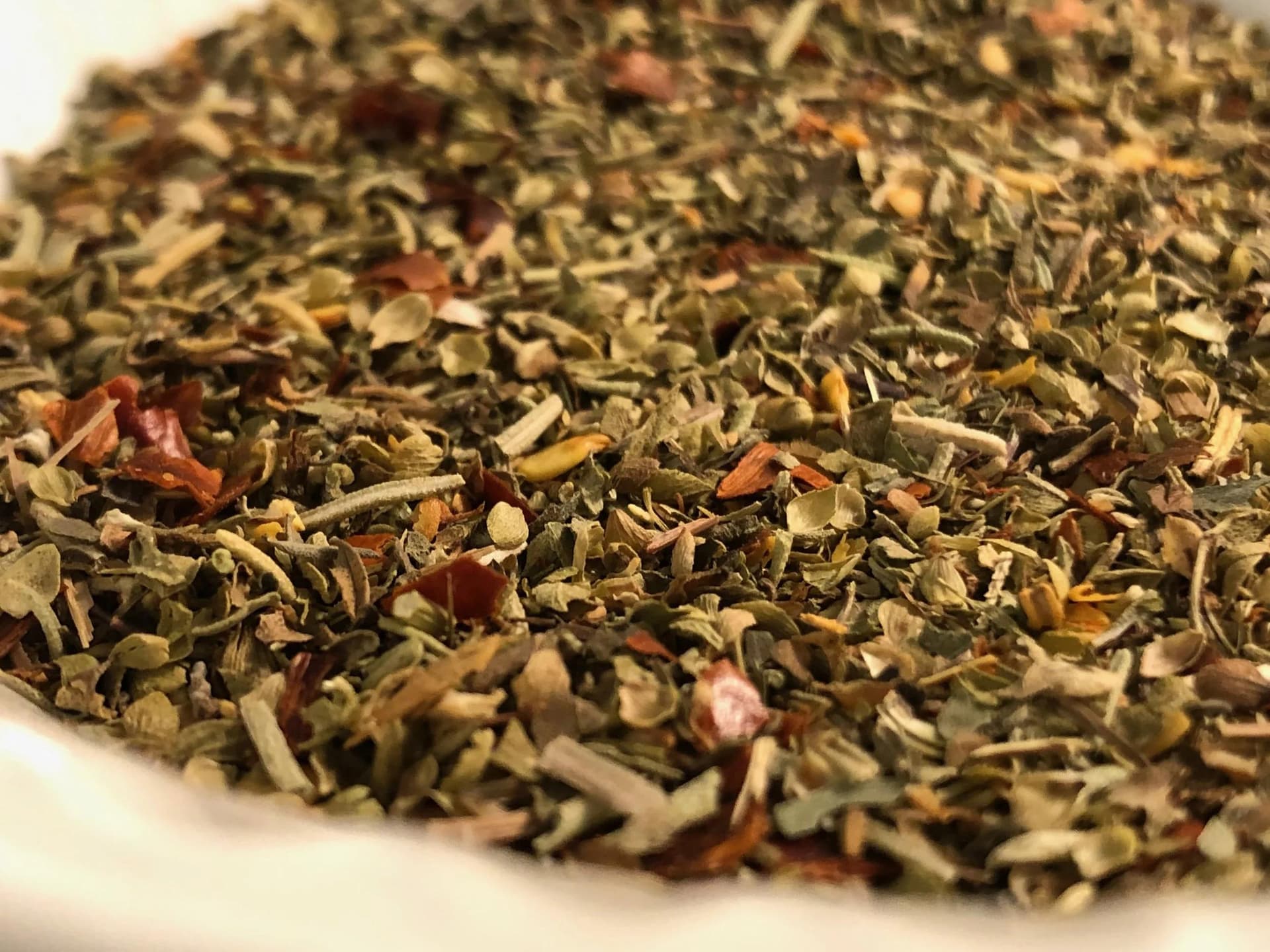 Close-up of homemade Italian seasoning blend with dried green herbs and red pepper flakes in a white bowl.