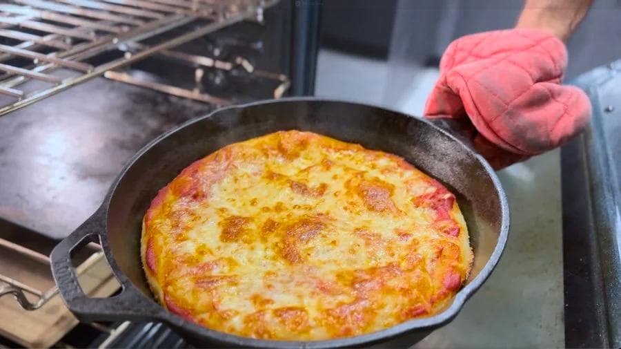 Cast iron skillet pizza with melted cheese and toppings being removed from oven with red oven mitts.